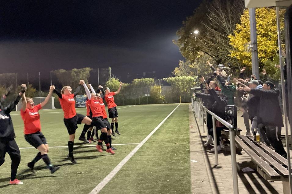 Feiernde Fu&szlig;ballmannschaft in roten Trikots jubelt auf dem Platz, umgeben von begeisterten Fans auf der Trib&uuml;ne.