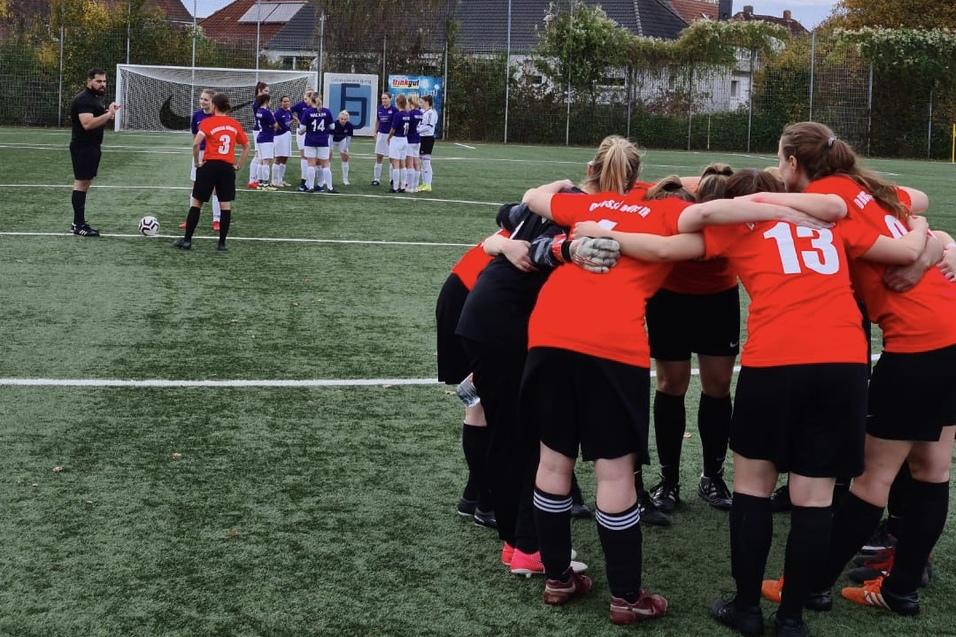Eine Gruppe von Fu&szlig;ballspielerinnen in roten Trikots bildet einen Kreis, w&auml;hrend eine gegnerische Mannschaft im Hintergrund steht.