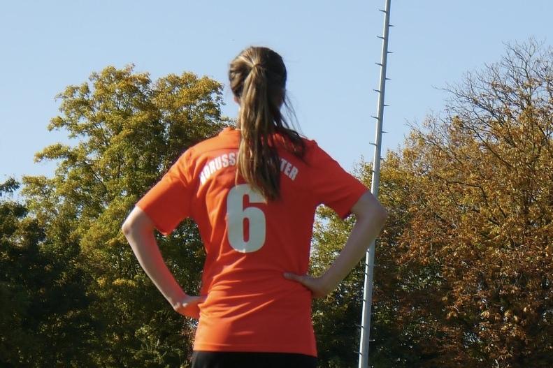 Eine Spielerin in einem orangefarbenen Trikot mit der Nummer 6 steht mit verschr&auml;nkten Armen auf einem Sportfeld.