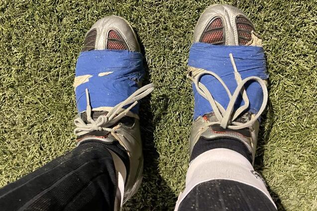 Nahaufnahme von F&uuml;&szlig;en in Fu&szlig;ballschuhen, mit blauer Klebebandreparatur, auf einem Kunstrasenplatz.