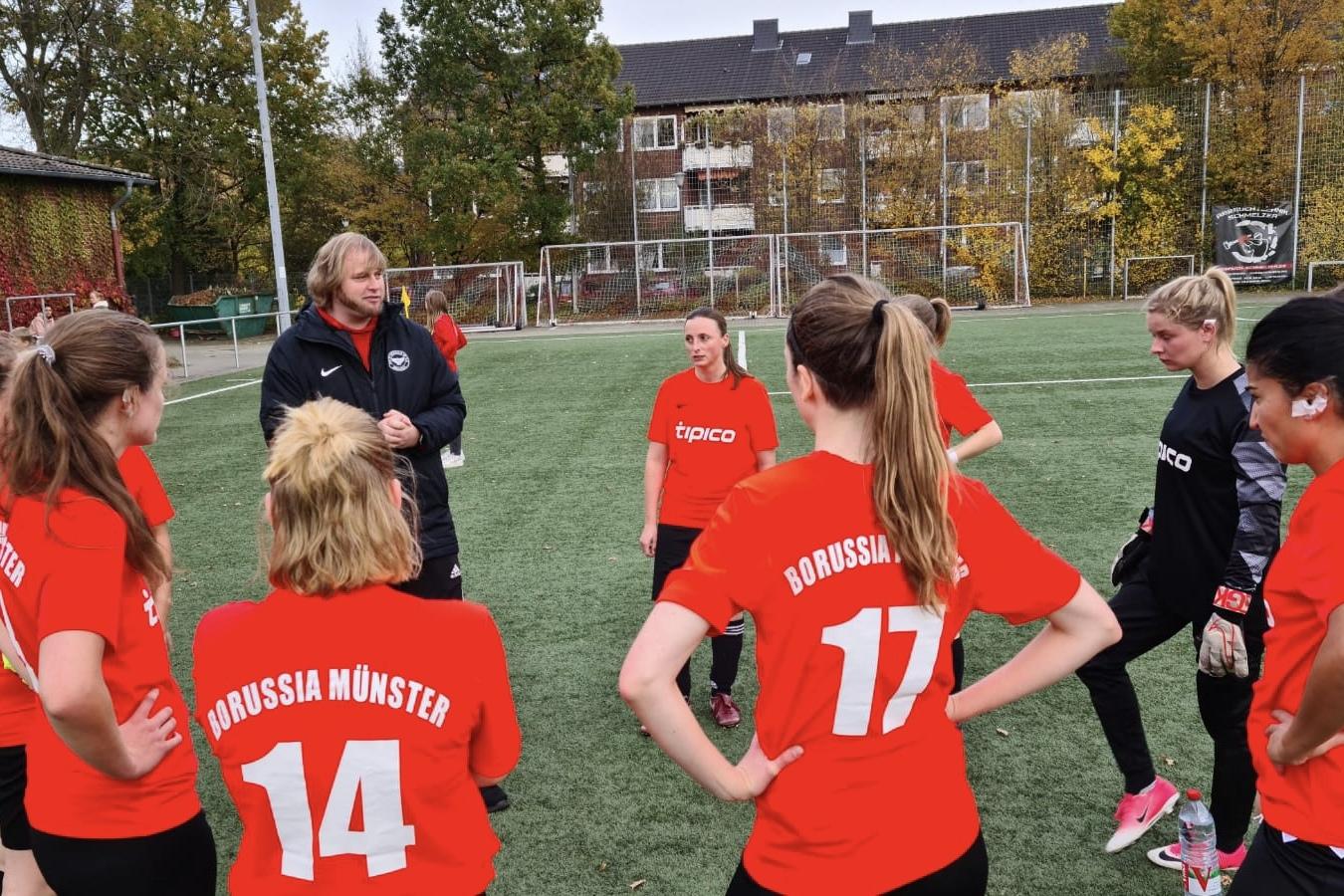 Trainer spricht mit einer Gruppe weiblicher Fu&szlig;ballspielerinnen in roten Trikots auf einem Rasenplatz.