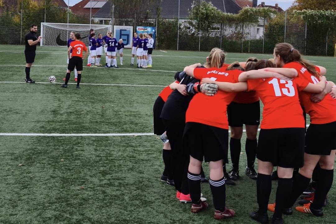 Eine Gruppe von Fu&szlig;ballspielerinnen in roten Trikots bildet einen Kreis, w&auml;hrend eine gegnerische Mannschaft im Hintergrund steht.