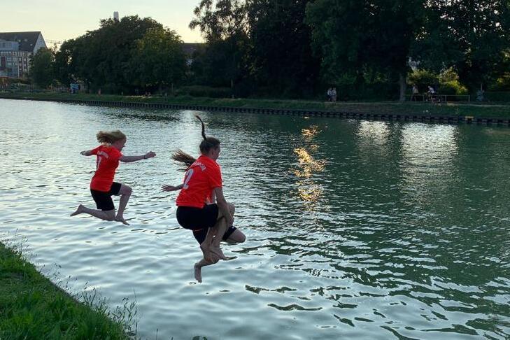 Zwei Kinder in roten T-Shirts springen in einen ruhigen Kanal bei Sonnenuntergang. Gr&uuml;ne Wiese im Vordergrund.