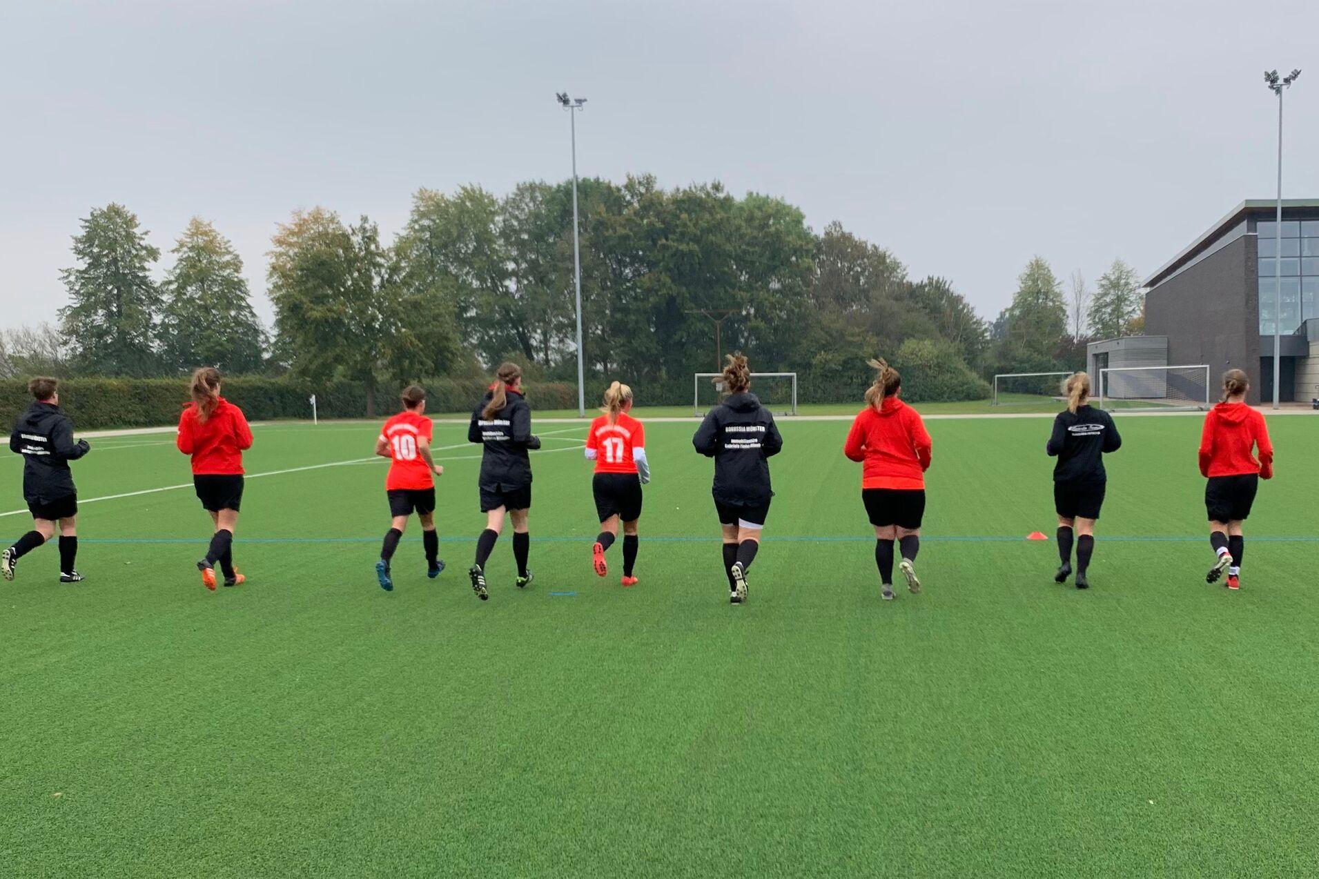 Eine Gruppe von zehn fu&szlig;ballspielenden Frauen joggt auf einem Kunstrasenplatz, umgeben von B&auml;umen und einem Sportgeb&auml;ude.