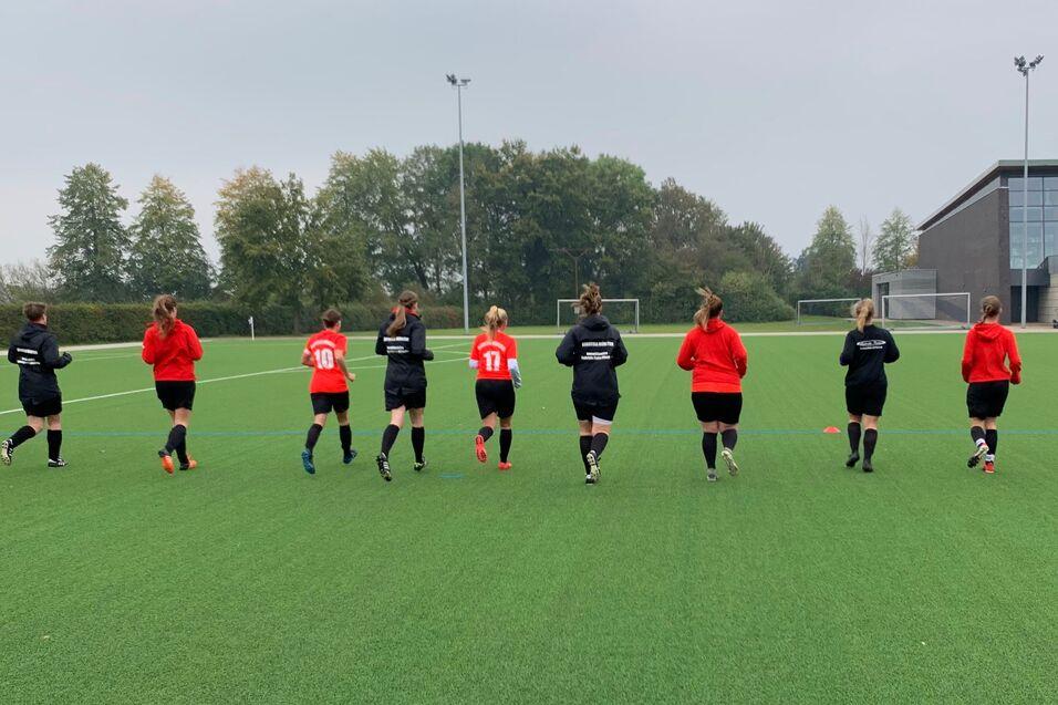 Eine Gruppe von zehn fu&szlig;ballspielenden Frauen joggt auf einem Kunstrasenplatz, umgeben von B&auml;umen und einem Sportgeb&auml;ude.