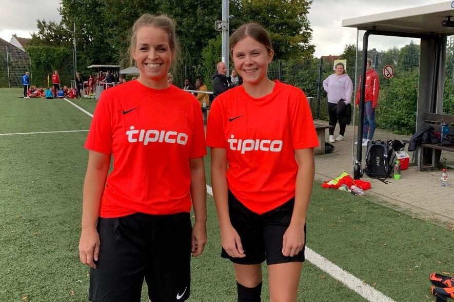 Zwei Spielerinnen in roten Trikots stehen auf einem Fu&szlig;ballplatz vor einer Zuschauertrib&uuml;ne.