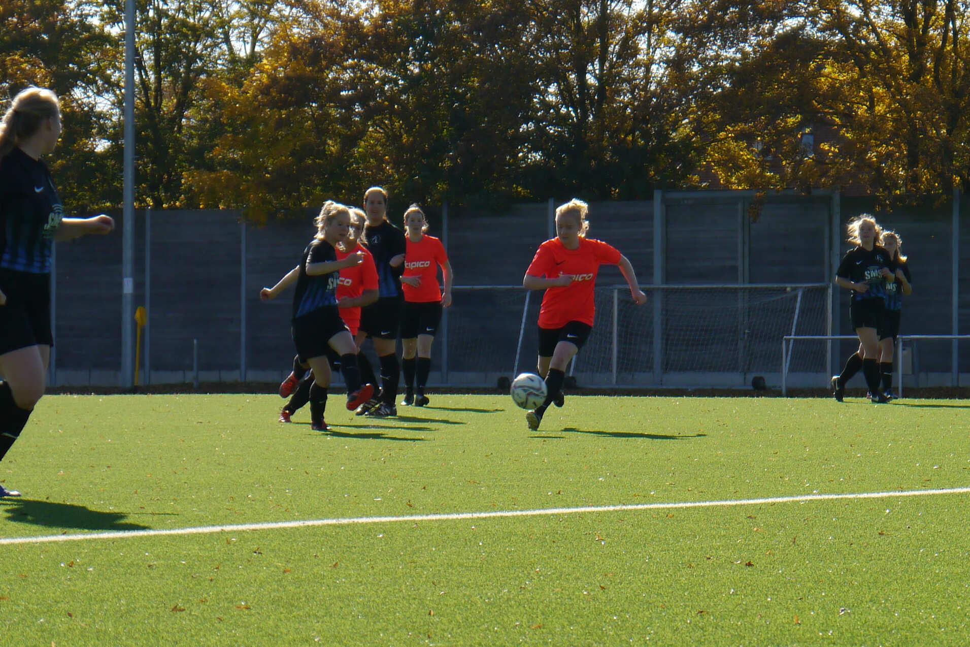 Spielerinnen in roten und schwarzen Trikots laufen &uuml;ber ein gr&uuml;nes Fu&szlig;ballfeld, w&auml;hrend eine Spielerin den Ball dribbelt.