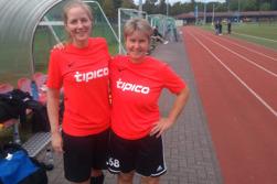 Zwei Fu&szlig;ballspielerinnen in roten Trikots stehen l&auml;chelnd neben einer Sportbank auf dem Platz.