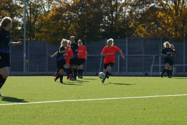 Spielerinnen in roten und schwarzen Trikots laufen &uuml;ber ein gr&uuml;nes Fu&szlig;ballfeld, w&auml;hrend eine Spielerin den Ball dribbelt.