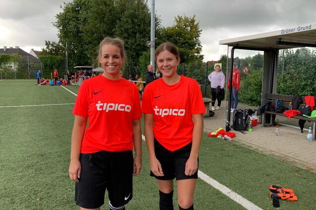 Zwei Spielerinnen in roten Trikots stehen auf einem Fu&szlig;ballplatz vor einer Zuschauertrib&uuml;ne.