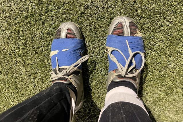 Nahaufnahme von F&uuml;&szlig;en in Fu&szlig;ballschuhen, mit blauer Klebebandreparatur, auf einem Kunstrasenplatz.