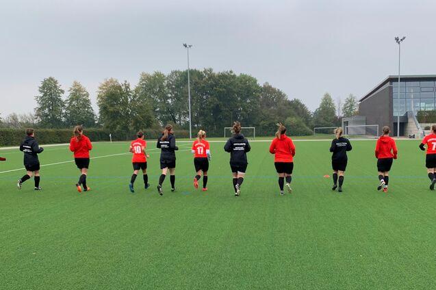 Eine Gruppe von zehn fu&szlig;ballspielenden Frauen joggt auf einem Kunstrasenplatz, umgeben von B&auml;umen und einem Sportgeb&auml;ude.