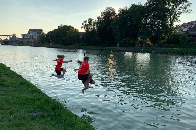 Zwei Kinder in roten T-Shirts springen in einen ruhigen Kanal bei Sonnenuntergang. Gr&uuml;ne Wiese im Vordergrund.