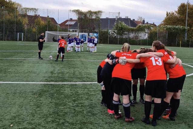 Eine Gruppe von Fu&szlig;ballspielerinnen in roten Trikots bildet einen Kreis, w&auml;hrend eine gegnerische Mannschaft im Hintergrund steht.
