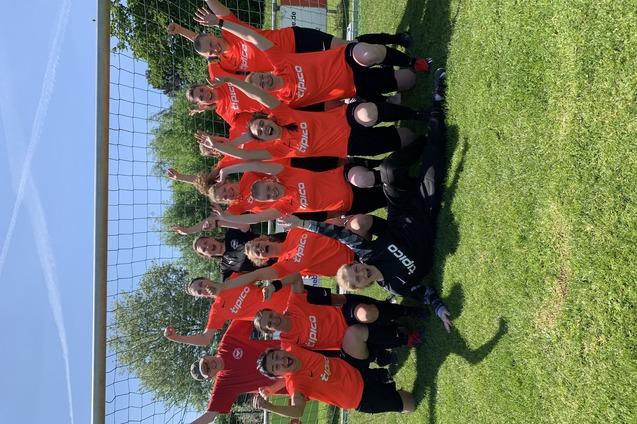 Gruppenfoto von 14 Fu&szlig;ballspielern in roten Trikots, die vor einem Tor auf einer Wiese jubeln und die Arme heben.