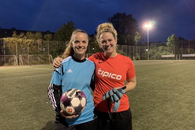 Zwei Frauen stehen l&auml;chelnd auf einem Fu&szlig;ballplatz, eine h&auml;lt einen Fu&szlig;ball, die andere tr&auml;gt Torehandschuhe.