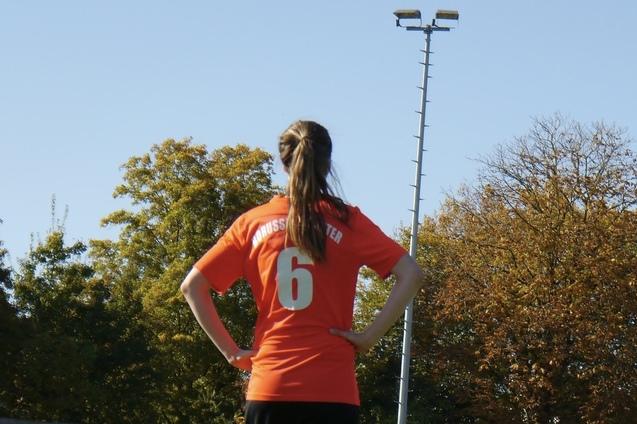 Eine Spielerin in einem orangefarbenen Trikot mit der Nummer 6 steht mit verschr&auml;nkten Armen auf einem Sportfeld.