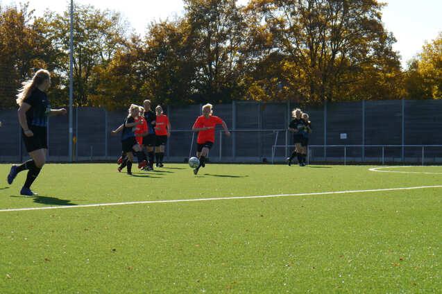 Spielerinnen in roten und schwarzen Trikots laufen &uuml;ber ein gr&uuml;nes Fu&szlig;ballfeld, w&auml;hrend eine Spielerin den Ball dribbelt.