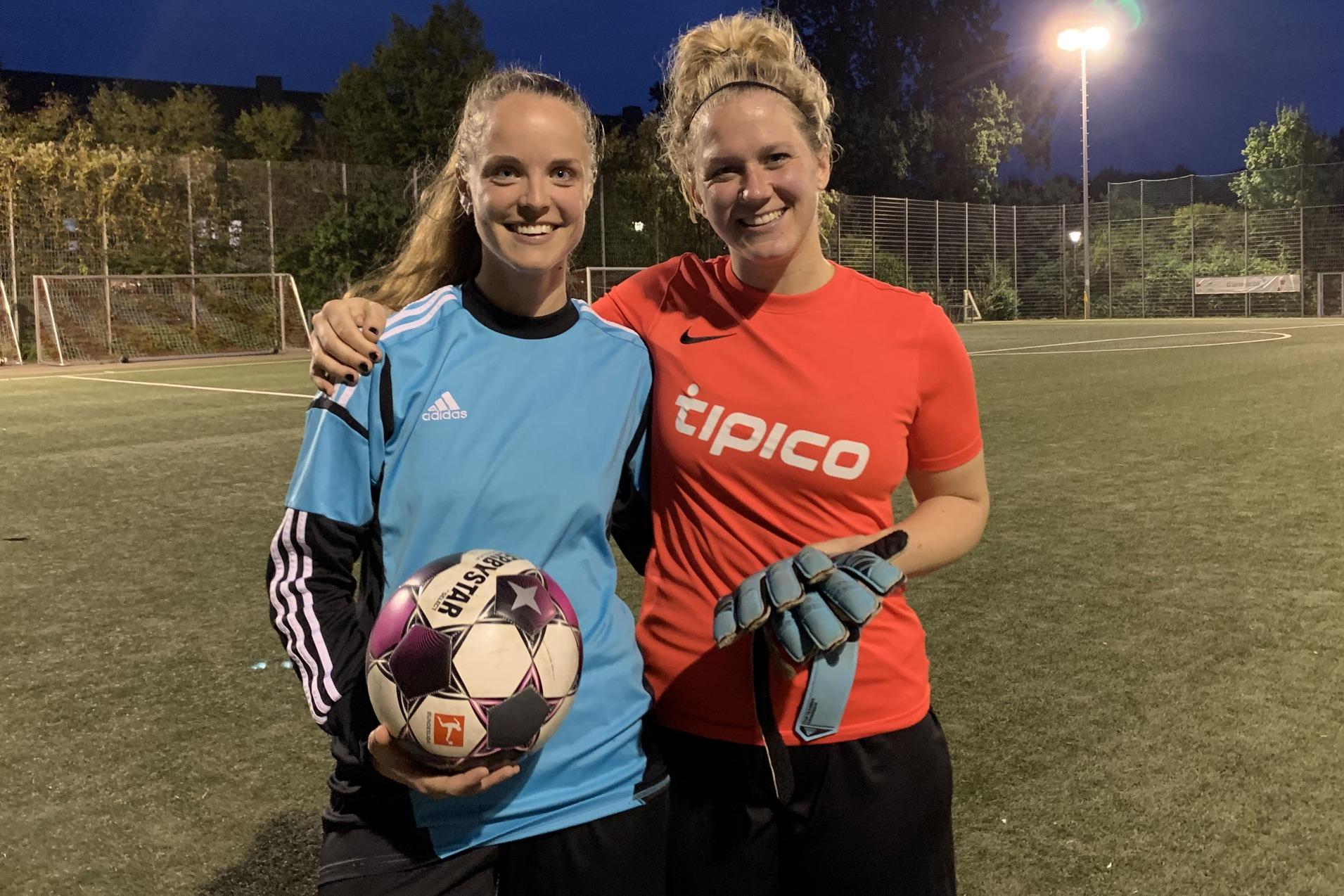 Zwei Frauen stehen l&auml;chelnd auf einem Fu&szlig;ballplatz, eine h&auml;lt einen Fu&szlig;ball, die andere tr&auml;gt Torehandschuhe.