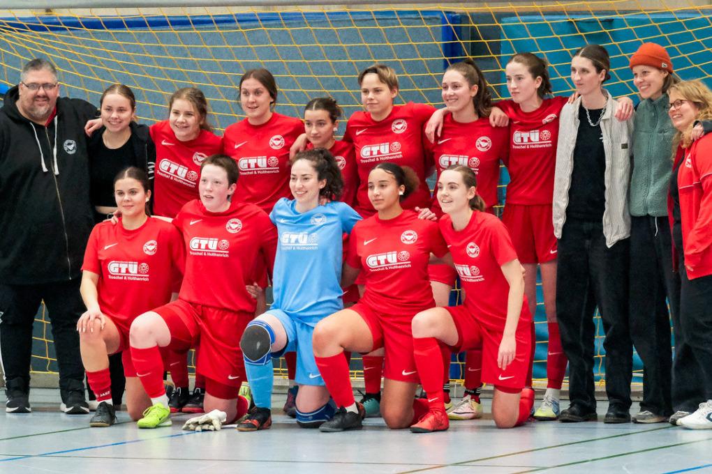 Mannschaftsfoto einer Fu&szlig;ballgruppe in roten Trikots mit Trainer, posierend vor einem Tor in einer Sporthalle.