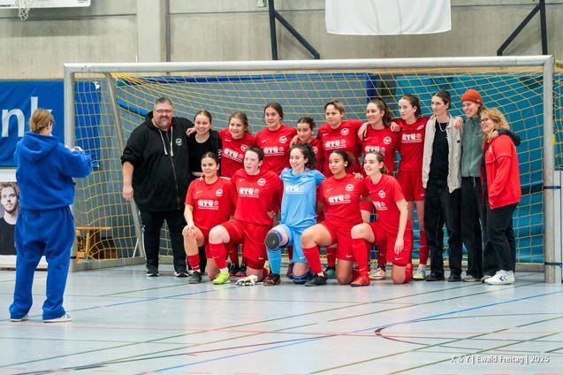 Mannschaftsfoto einer Fu&szlig;ballgruppe in roten Trikots mit Trainer, posierend vor einem Tor in einer Sporthalle.