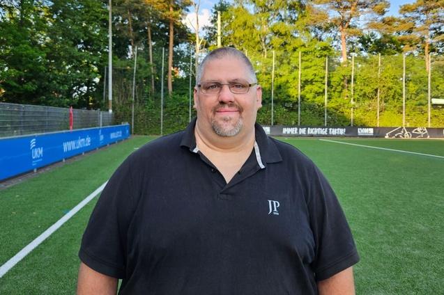 Mann mit Brille steht auf einem Sportplatz, im Hintergrund sind B&auml;ume und Fu&szlig;ballfeldmarkierungen zu sehen.