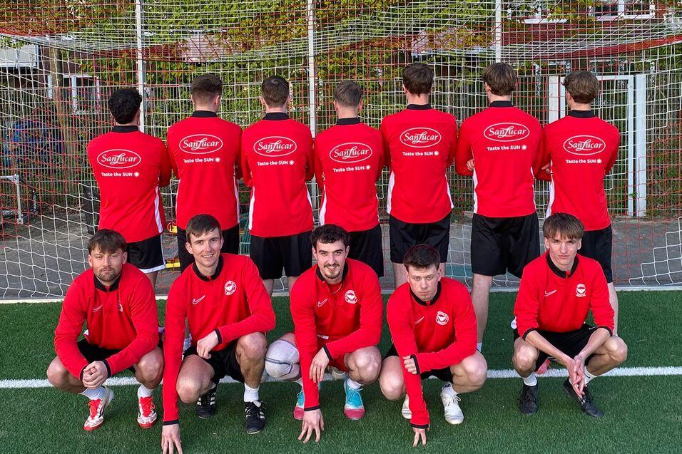 Gruppenfoto von zw&ouml;lf Fu&szlig;ballspielern in roten Trikots, verteilt vor und hinter einem Tor auf einem Rasenplatz.