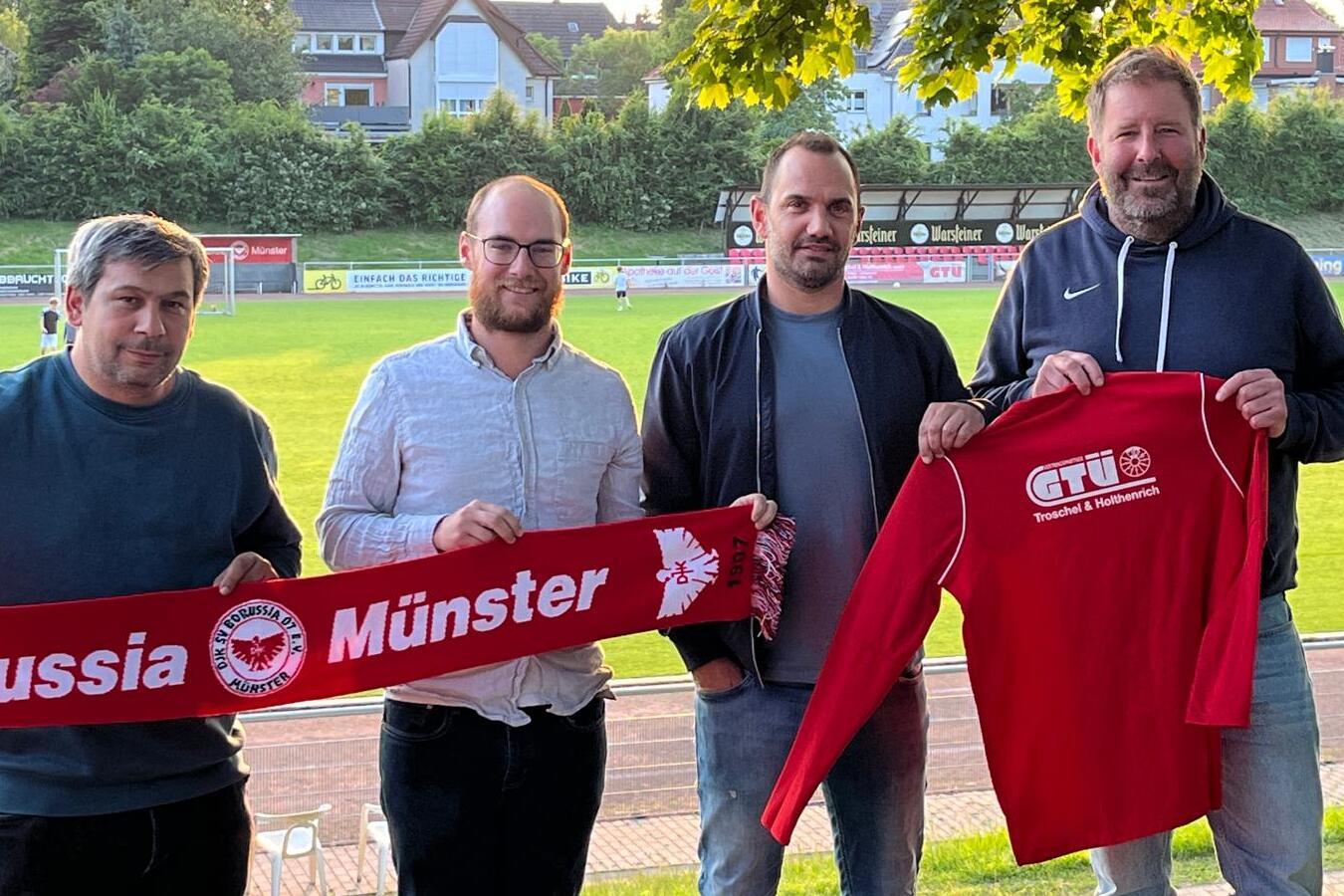 Vier M&auml;nner stehen am Spielfeldrand und halten ein Banner mit dem Schriftzug &bdquo;Borussia M&uuml;nster&ldquo; und ein rotes Trikot.