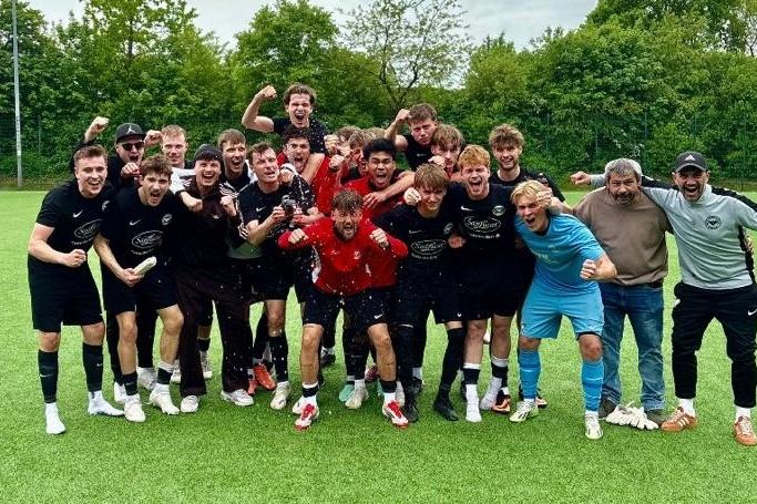 Gruppenshot eines Fu&szlig;ballteams in schwarzen Trikots, jubelnd auf dem Feld, mit einem Pokal in der Mitte.