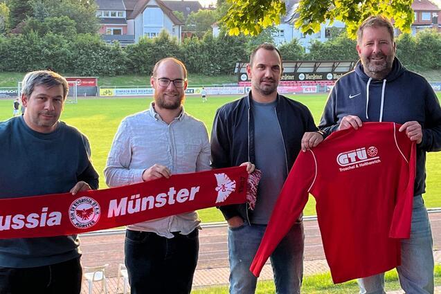 Vier M&auml;nner stehen am Spielfeldrand und halten ein Banner mit dem Schriftzug &bdquo;Borussia M&uuml;nster&ldquo; und ein rotes Trikot.