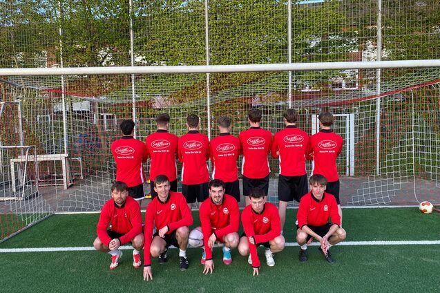 Gruppenfoto von zw&ouml;lf Fu&szlig;ballspielern in roten Trikots, verteilt vor und hinter einem Tor auf einem Rasenplatz.