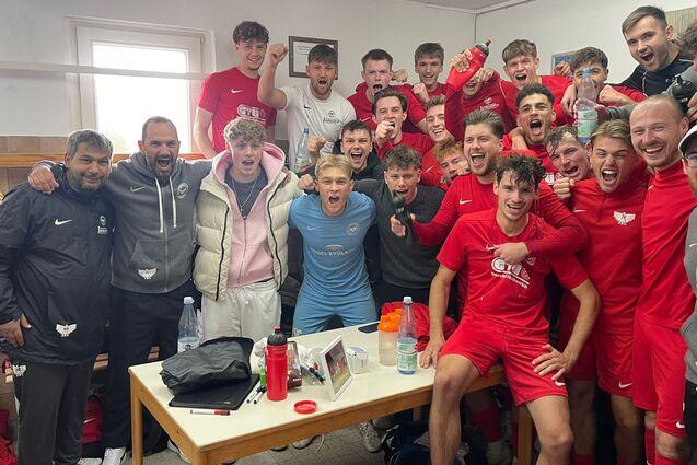 Ein begeistertes Fu&szlig;ballteam in roten Trikots versammelt sich in einem Umkleideraum, l&auml;chelnd und jubelnd.