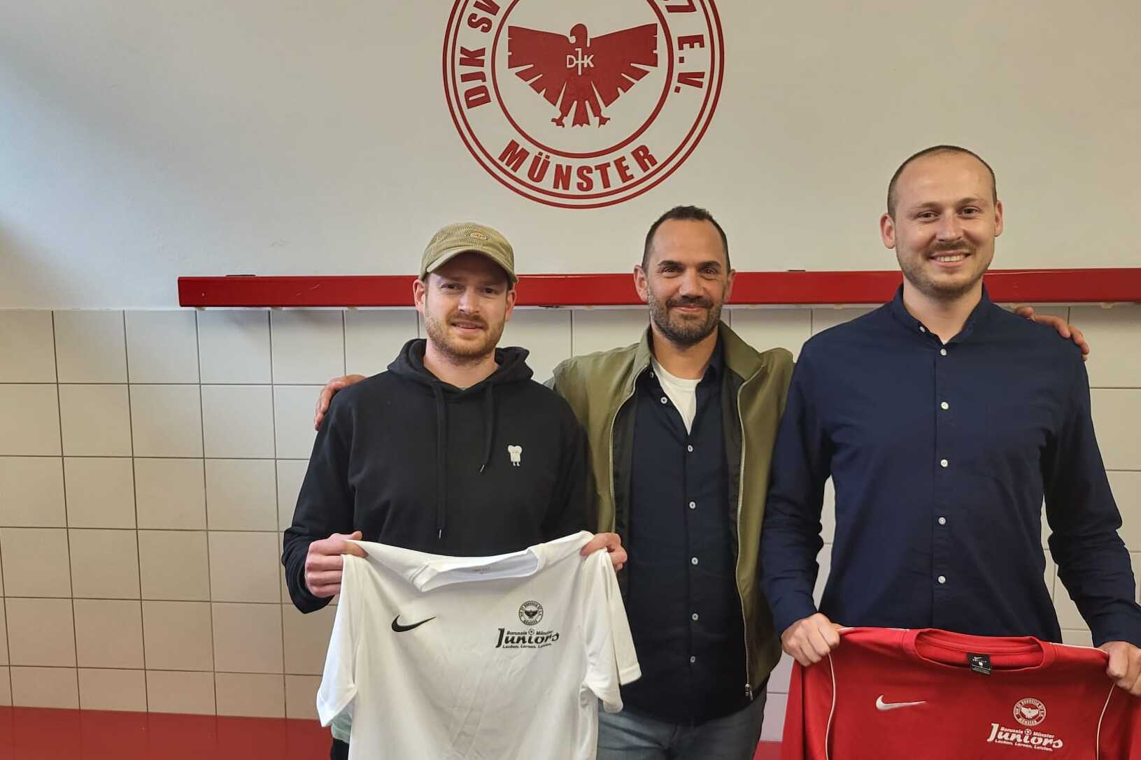 Drei M&auml;nner stehen in einem Umkleideraum und halten Fu&szlig;balltrikots eines Vereins mit Logo im Hintergrund.
