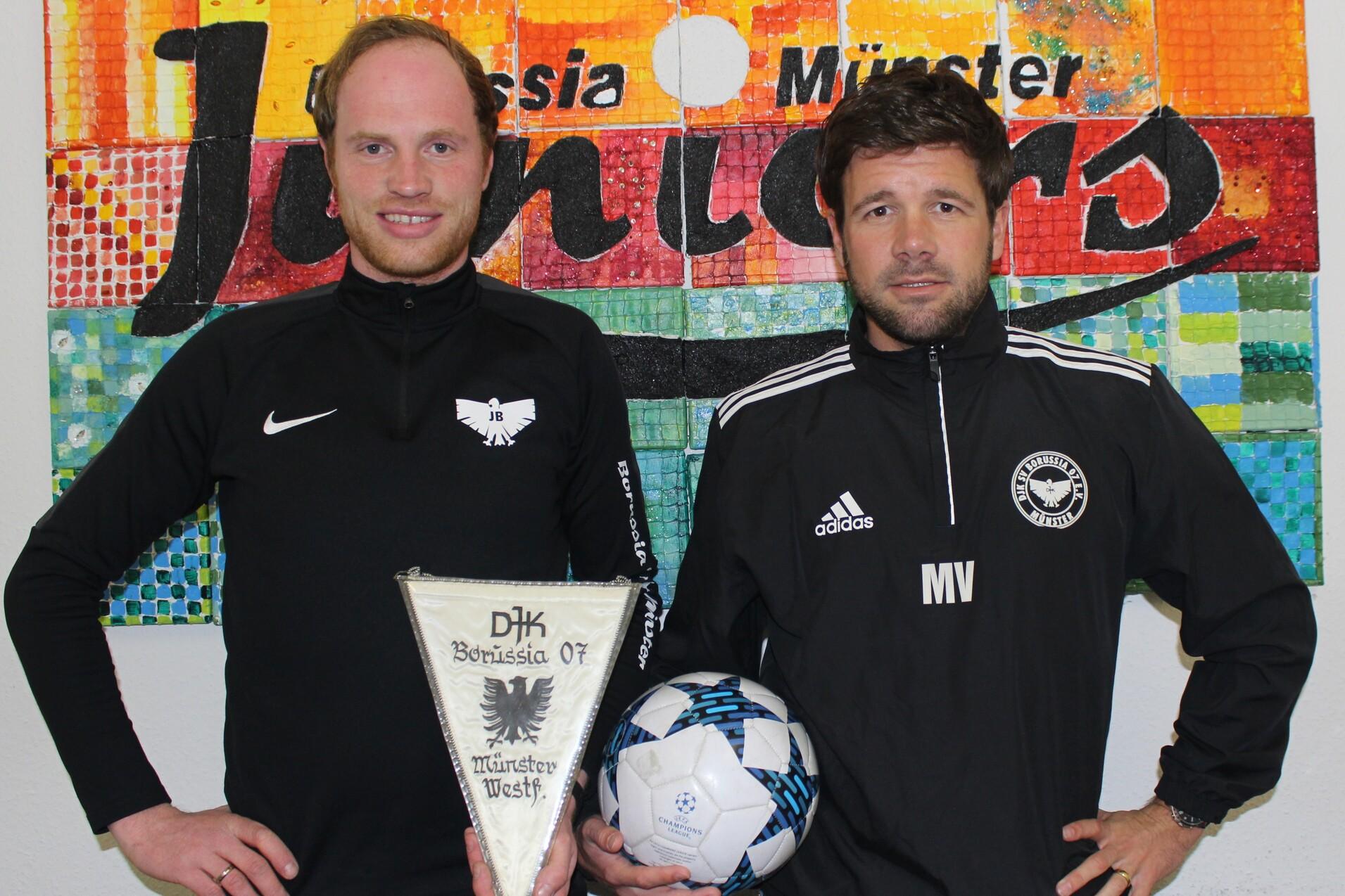 Zwei M&auml;nner mit Fu&szlig;ballbekleidung stehen vor einer farbigen Wand. Einer h&auml;lt einen Pokal, der andere einen Fu&szlig;ball.
