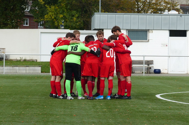 Eine Gruppe von Fu&szlig;ballspielern in roten Trikots bildet einen engen Kreis auf einem Rasenplatz.