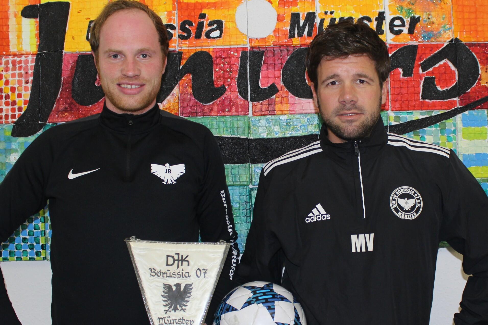 Zwei Männer mit Fußballbekleidung stehen vor einer farbigen Wand. Einer hält einen Pokal, der andere einen Fußball.