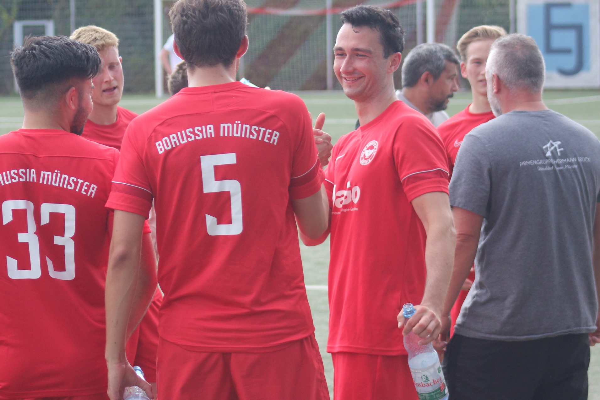 Gruppe von Fu&szlig;ballspielern in roten Trikots mit &bdquo;Borussia M&uuml;nster&ldquo;-Aufdruck beim Teamgespr&auml;ch auf dem Platz.