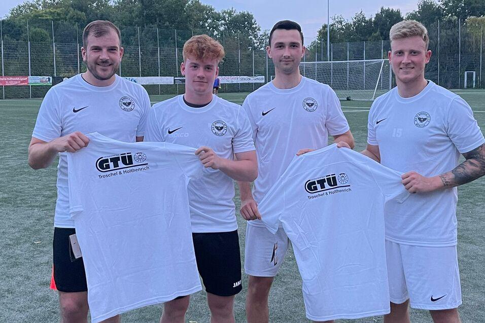 Vier Fu&szlig;ballspieler in wei&szlig;en Trikots halten T-Shirts mit dem Logo eines Sponsors auf einem Sportplatz.