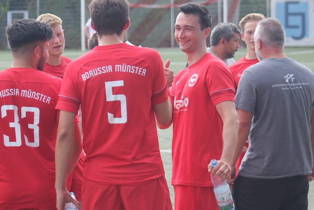 Gruppe von Fu&szlig;ballspielern in roten Trikots mit &bdquo;Borussia M&uuml;nster&ldquo;-Aufdruck beim Teamgespr&auml;ch auf dem Platz.