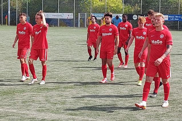 Acht Fu&szlig;ballspieler in roten Trikots gehen auf einem Kunstrasenplatz. Hintergrund sichtbar, mit Banden und B&auml;umen.
