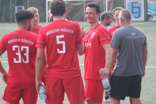 Gruppe von Fu&szlig;ballspielern in roten Trikots mit &bdquo;Borussia M&uuml;nster&ldquo;-Aufdruck beim Teamgespr&auml;ch auf dem Platz.