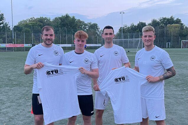 Vier Fu&szlig;ballspieler in wei&szlig;en Trikots halten T-Shirts mit dem Logo eines Sponsors auf einem Sportplatz.
