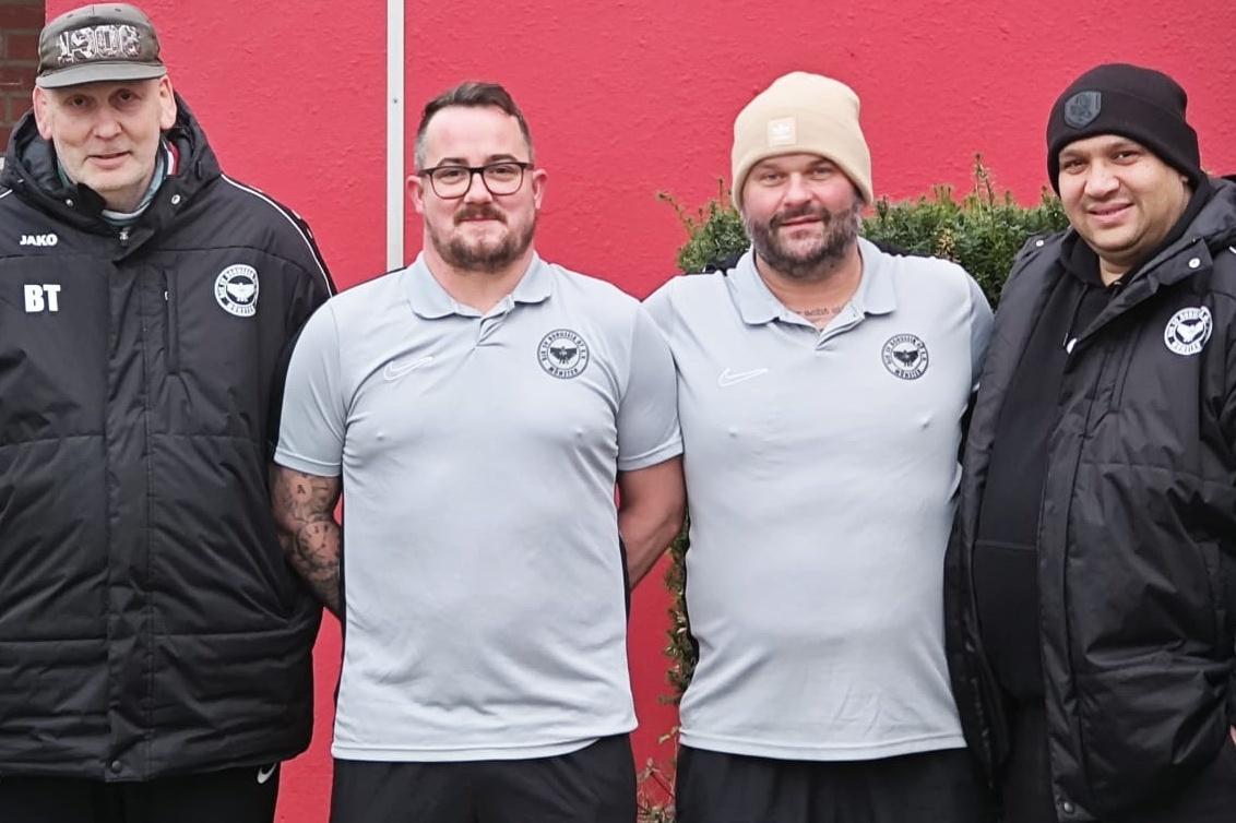 Vier M&auml;nner in Sportkleidung stehen vor gr&uuml;ner Hecke und roter Wand, alle l&auml;cheln in die Kamera.