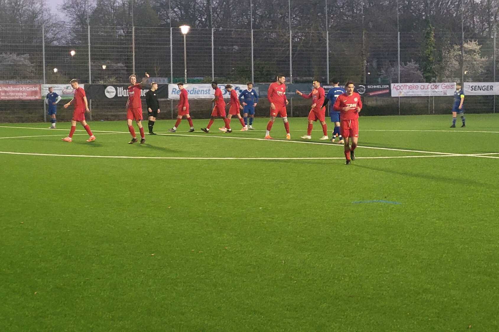 Fu&szlig;ballspiel auf Kunstrasen mit Spielern in roten und blauen Trikots, umgeben von einer n&auml;chtlichen Umgebung.