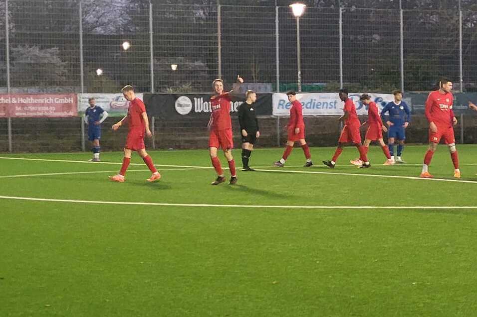 Fu&szlig;ballspiel auf Kunstrasen mit Spielern in roten und blauen Trikots, umgeben von einer n&auml;chtlichen Umgebung.
