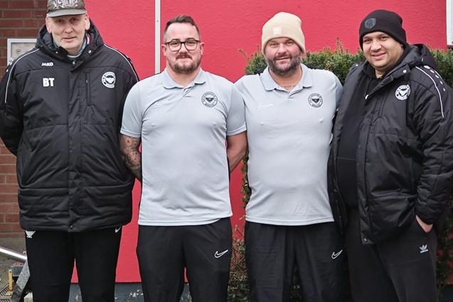 Vier M&auml;nner in Sportkleidung stehen vor gr&uuml;ner Hecke und roter Wand, alle l&auml;cheln in die Kamera.