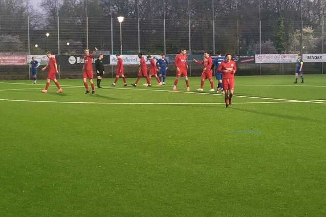 Fu&szlig;ballspiel auf Kunstrasen mit Spielern in roten und blauen Trikots, umgeben von einer n&auml;chtlichen Umgebung.