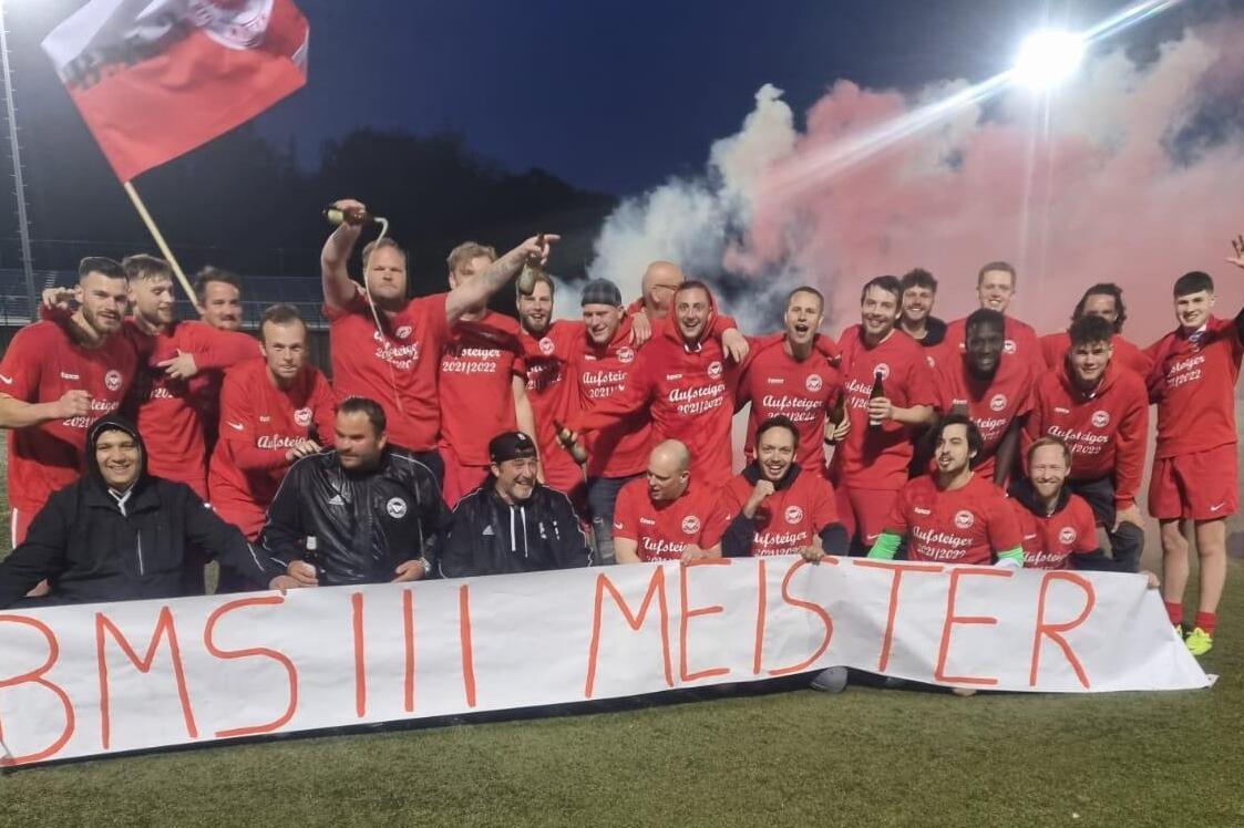 Gruppe von Spielern in roten Trikots posiert in der Nacht mit einem Banner &bdquo;BMS III Meister&ldquo; und Feuern im Hintergrund.
