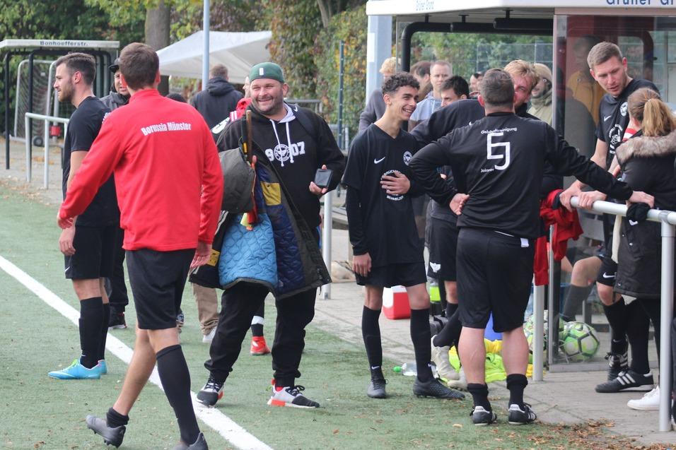 Spieler einer Fu&szlig;ballmannschaft stehen an der Bank, einige lachen und unterhalten sich, w&auml;hrend B&auml;lle im Hintergrund liegen.