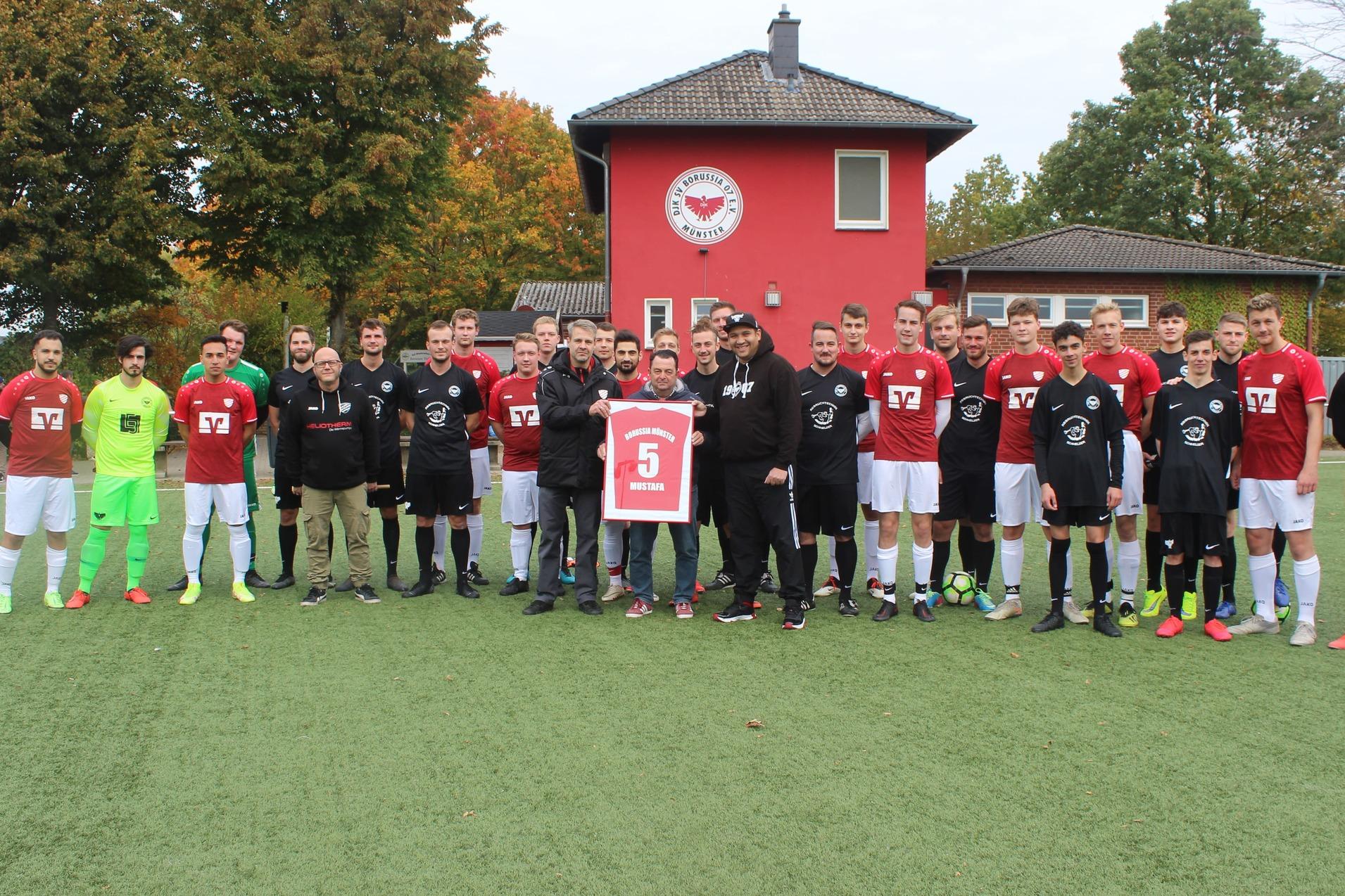 Gruppenfoto von zwei Fu&szlig;ballmannschaften in roten und schwarzen Trikots, stehend auf einem Kunstrasenplatz vor einem Vereinsgeb&auml;ude.